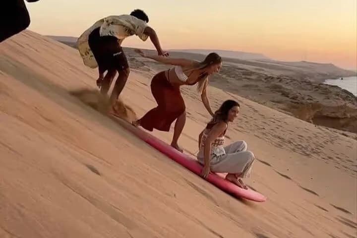 Friends running down a sand dune with a sandboard at sunset